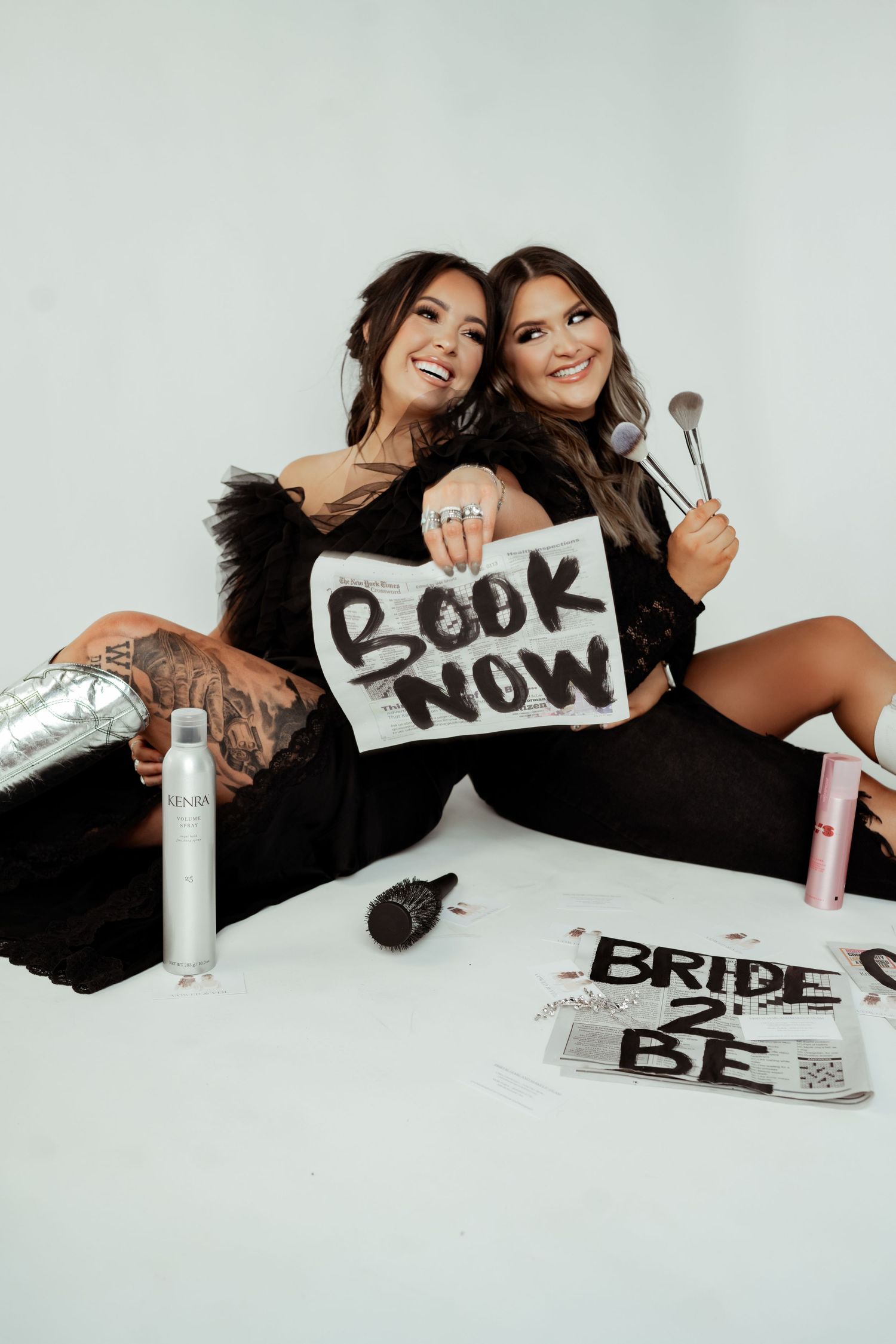 Two women in black outfits pose with makeup brushes and a Book Now sign, surrounded by beauty products and a Bride 2 Be sign.