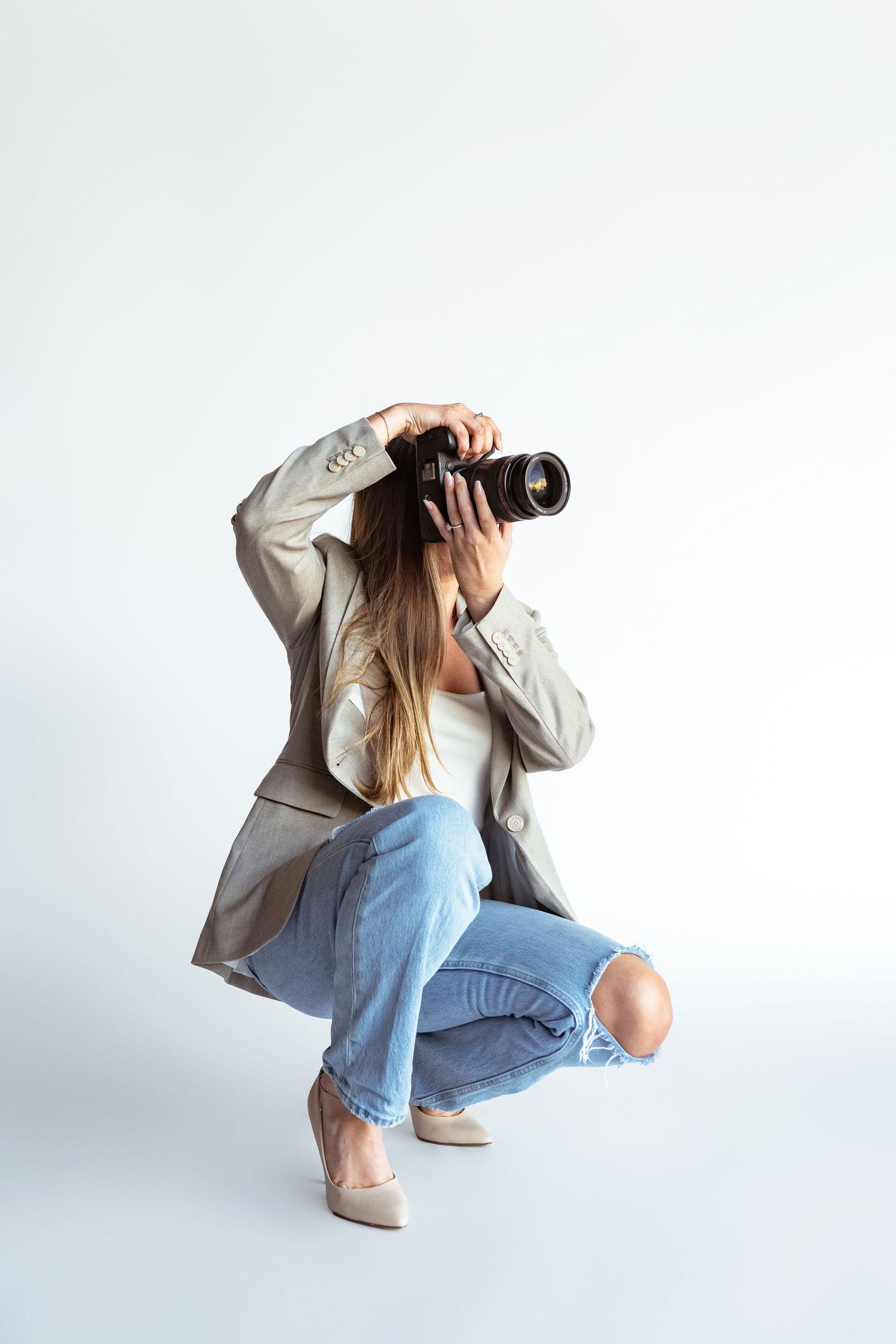 Woman crouching in ripped jeans and blazer, holding a professional DSLR camera up to her face on white background.