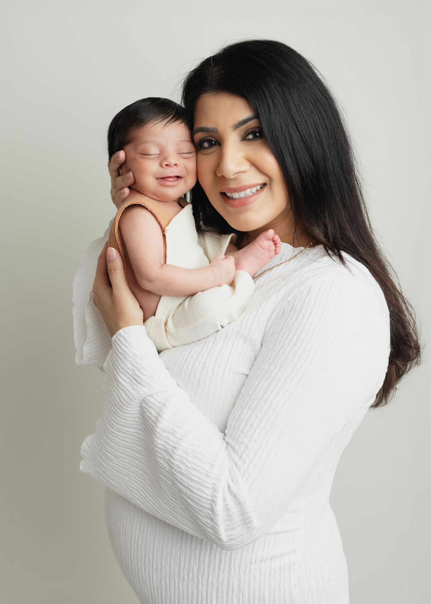 A smiling mother in a white ribbed dress holds her sleeping newborn baby against a soft neutral background. Elizabeth Lang Photography Surre