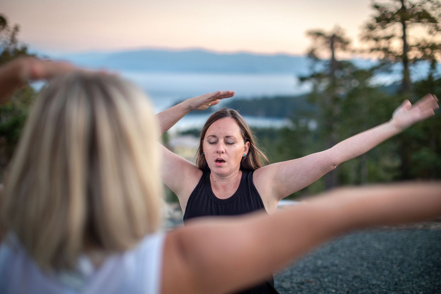 Two women practicing yoga outdoors at sunset with scenic lake and forest views.