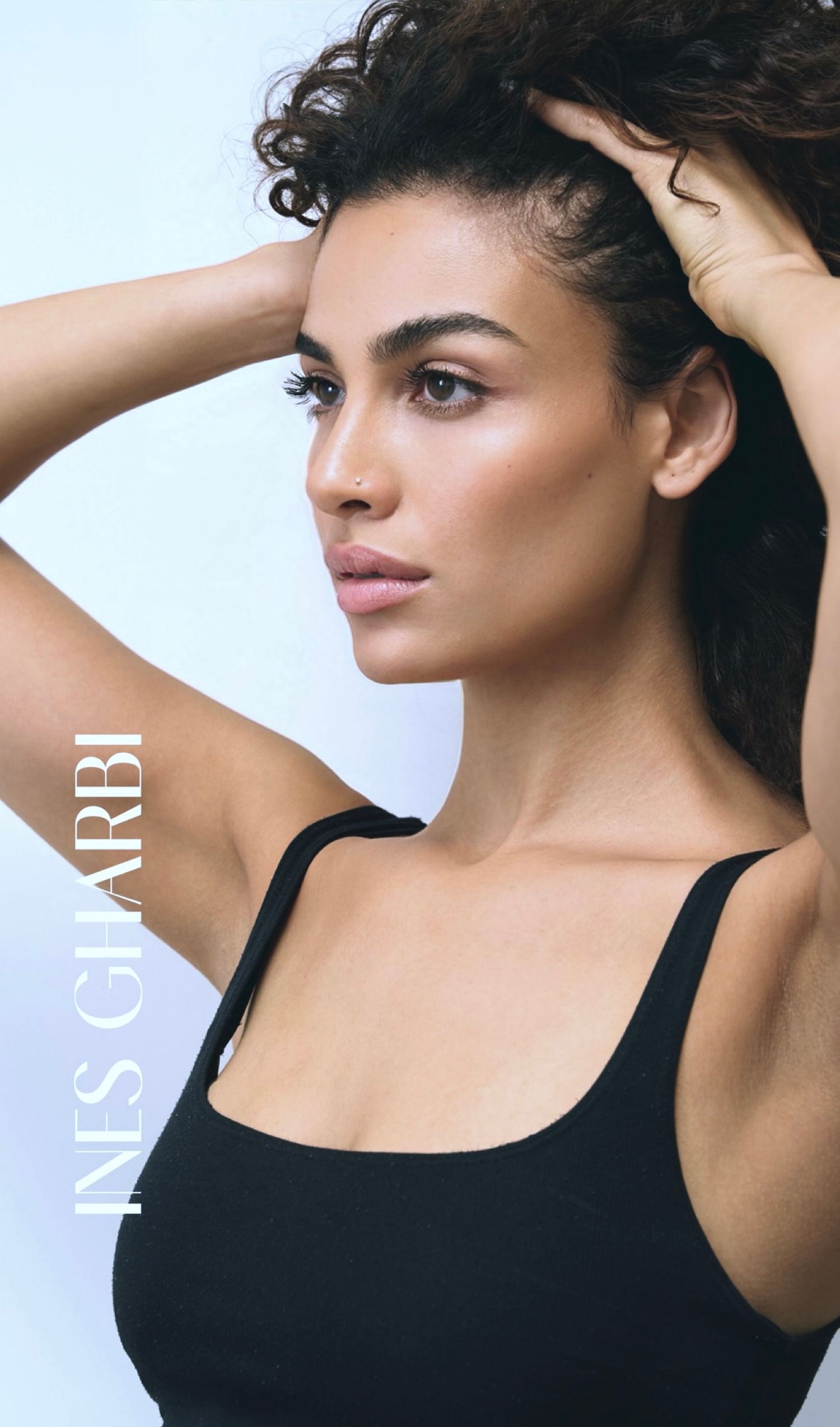 Ines Gharbi poses in a black tank top with hands raised, showcasing natural beauty and glowing skin against a light background.