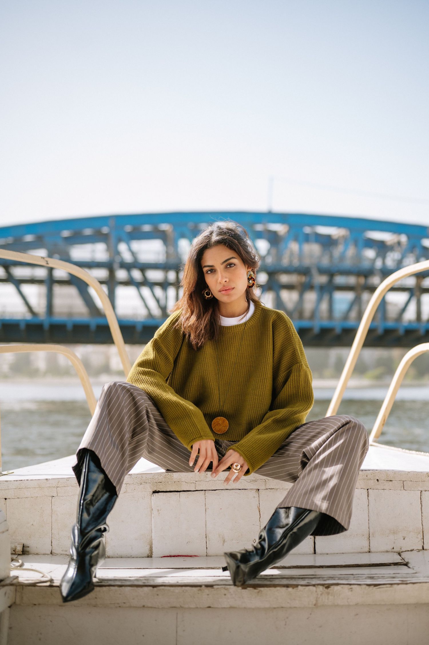 Woman in olive sweater and striped pants sits on a boat with a blue bridge in the background.