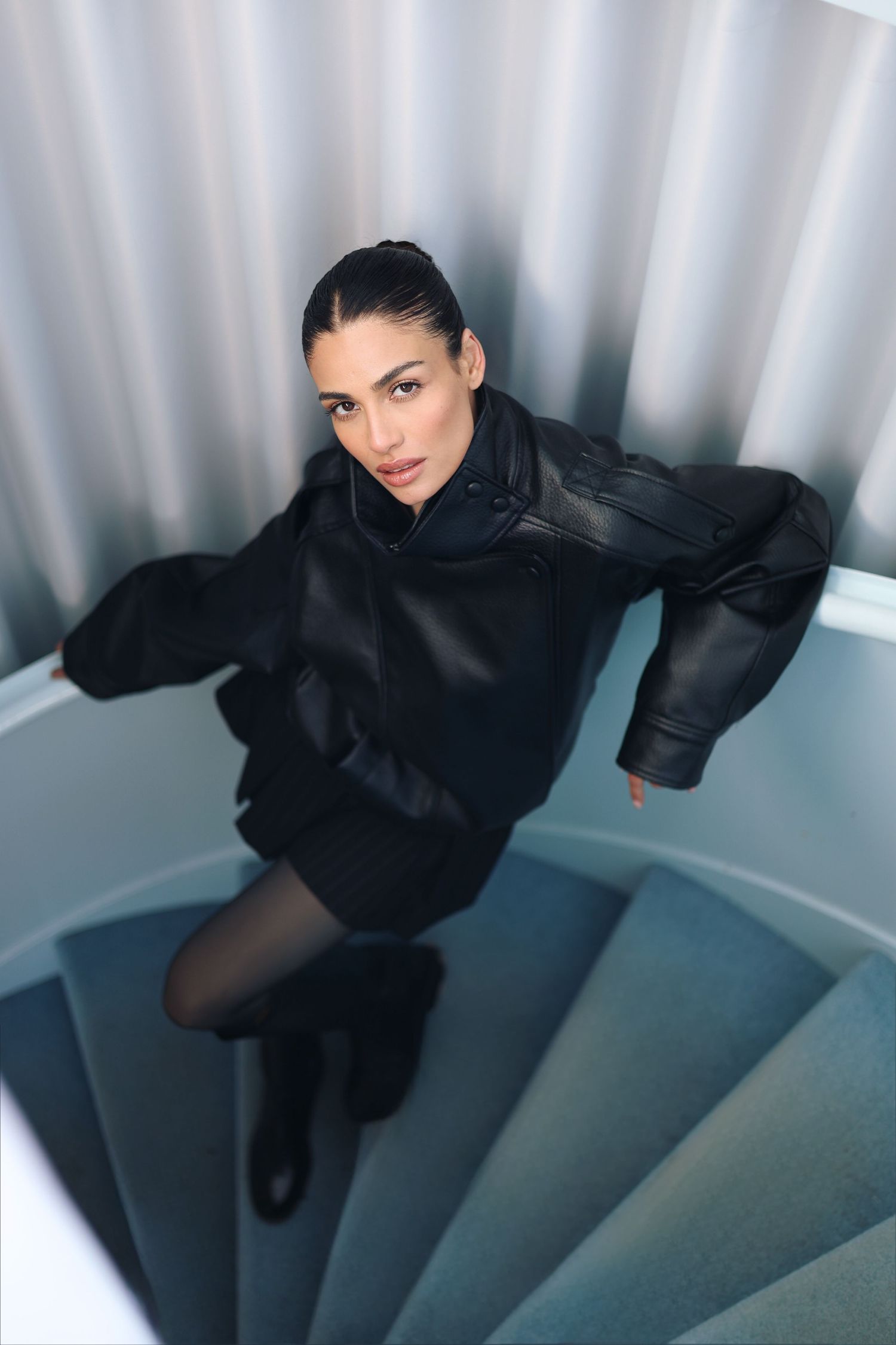 Woman in black leather jacket posing on spiral staircase, looking up at camera with modern metallic background.