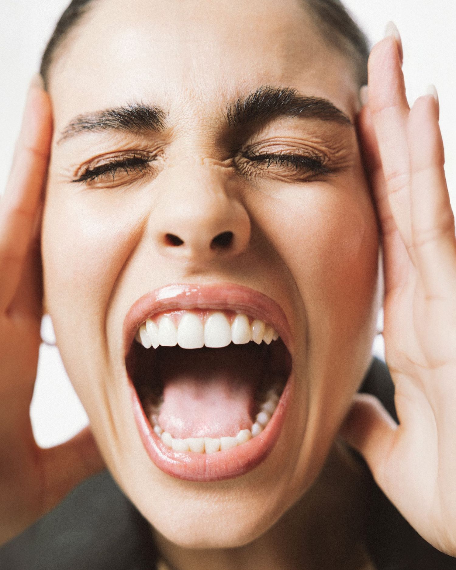 Close-up of a woman screaming with hands on her face, eyes shut, expressing intense emotion or stress.