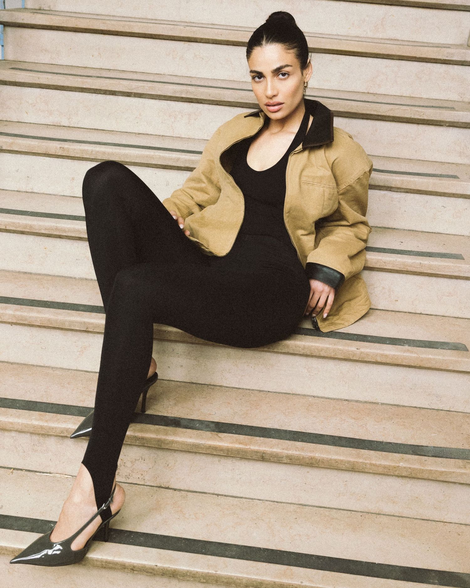 Woman in tan jacket, black bodysuit and leggings, silver heels, sitting on marble steps in a stylish pose.