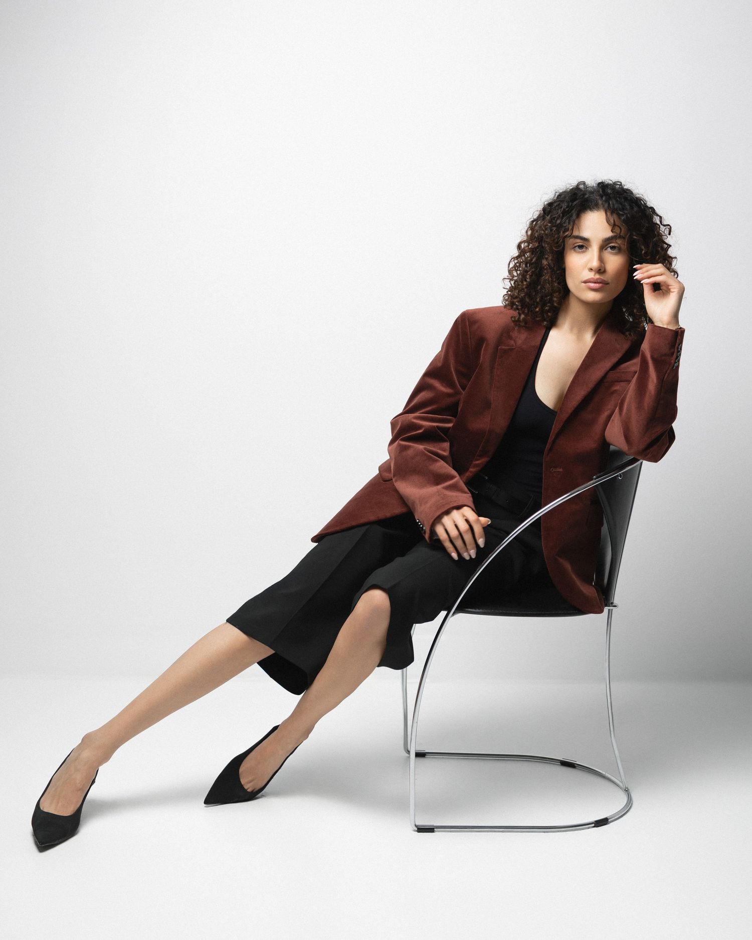 Woman in burgundy blazer and black dress sits elegantly on modern chrome chair against white background.