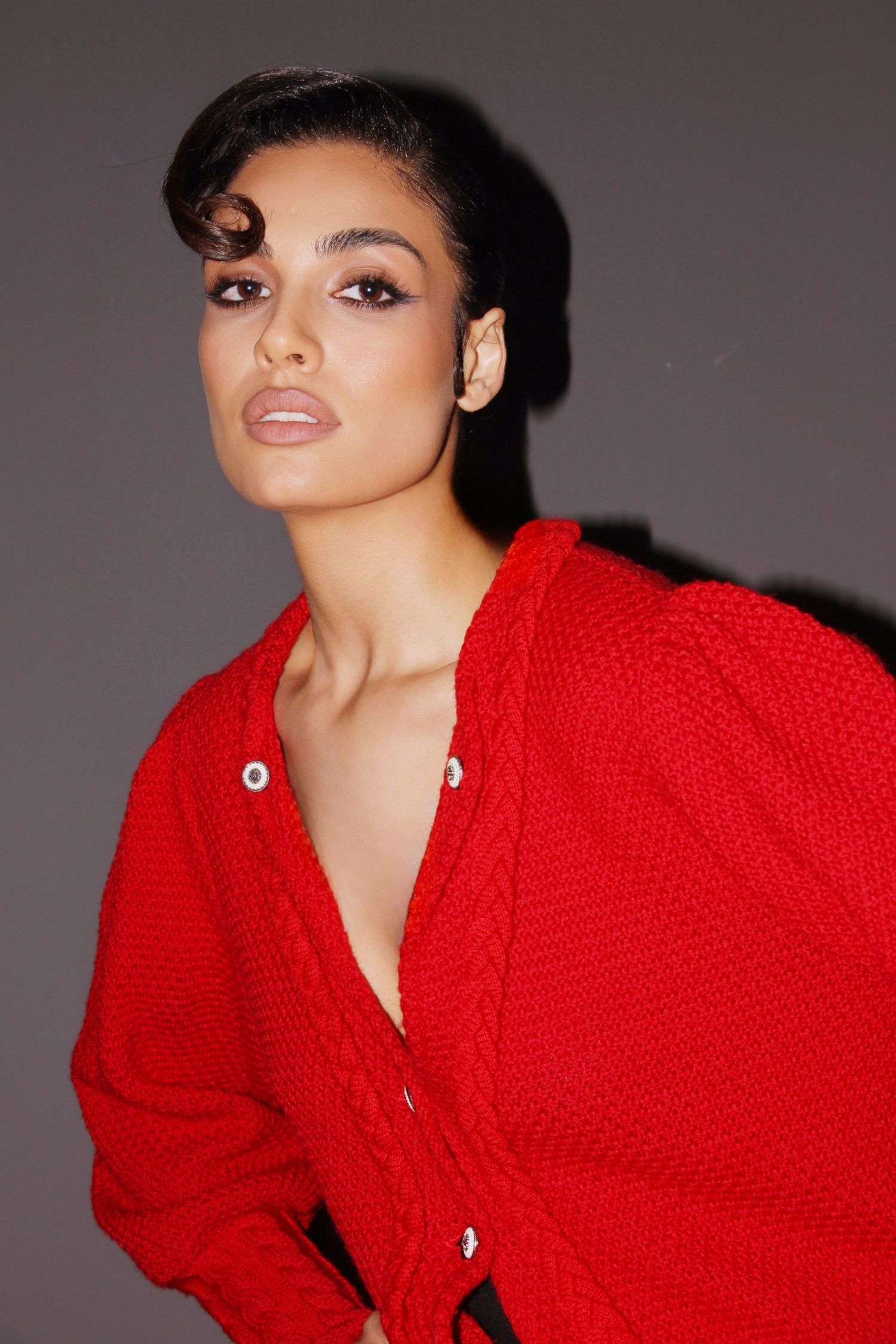 A woman in a red cable-knit cardigan poses confidently against a gray background with dramatic makeup.