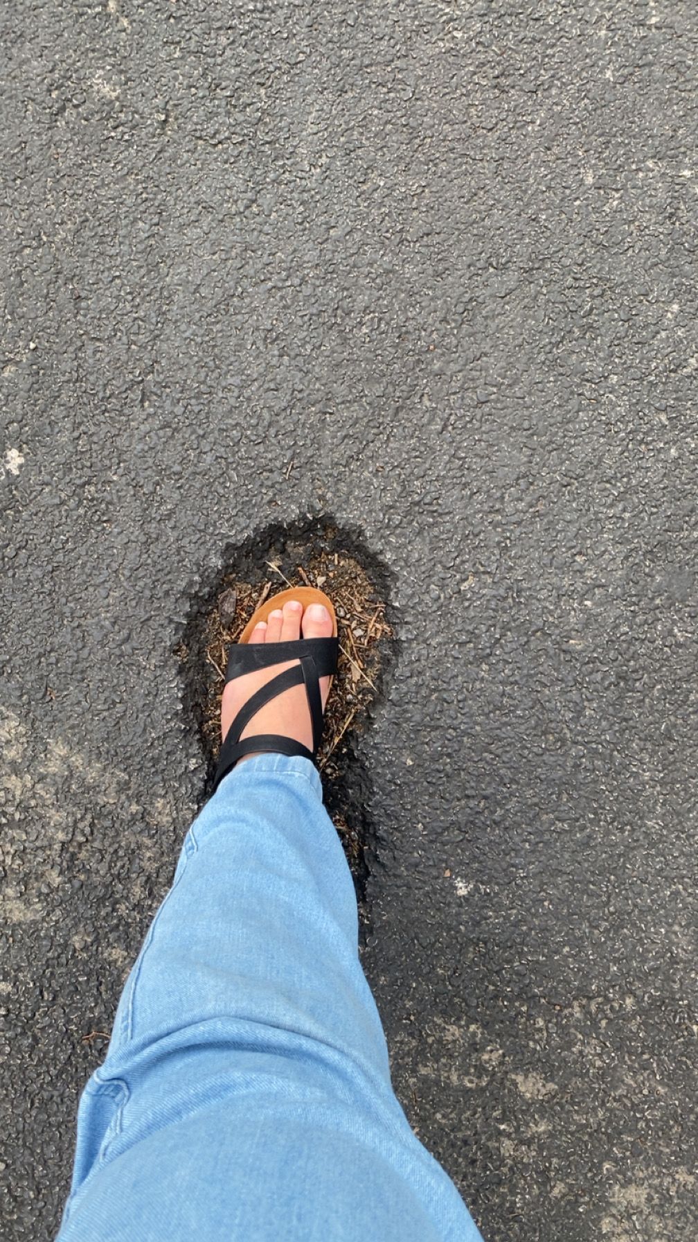 Person wearing black strappy sandals and blue jeans standing on asphalt with scattered debris underfoot.