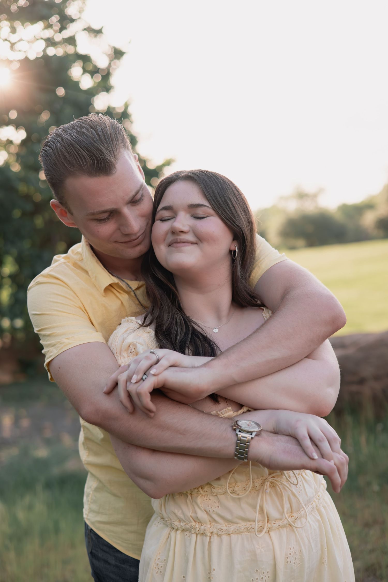 Couple embracing outdoors at golden hour, man in yellow polo hugging woman in yellow dress, smiling with eyes closed.