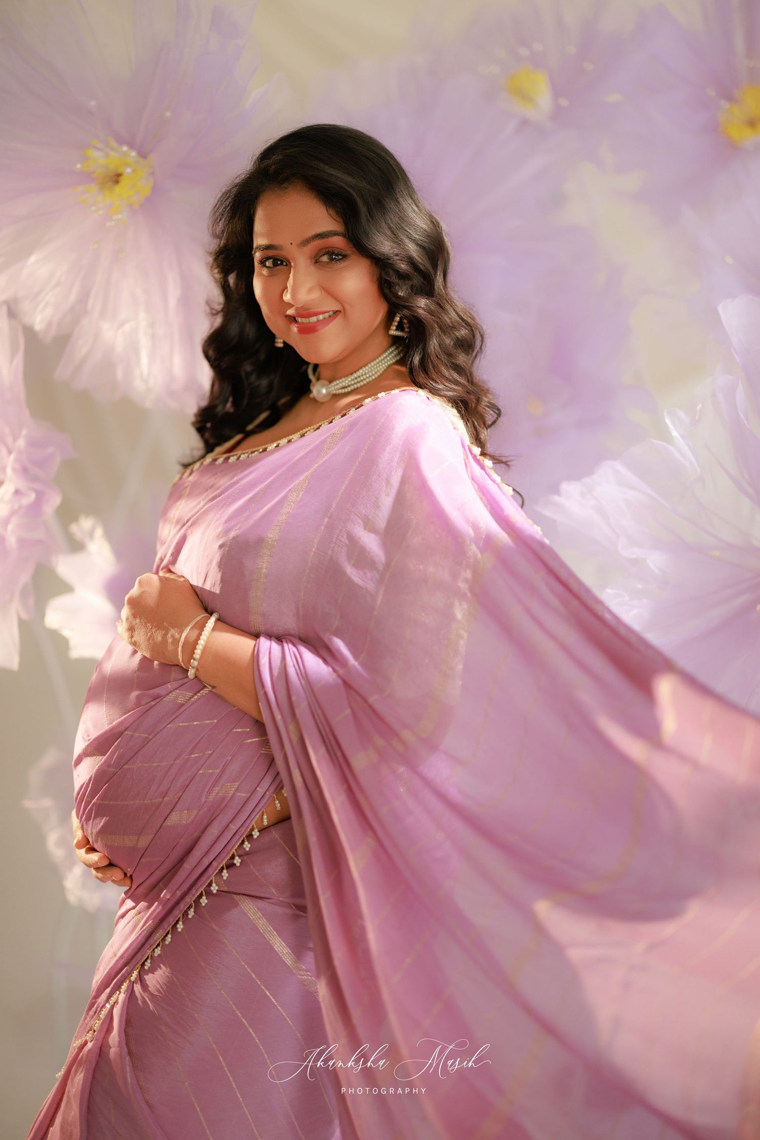 A person in a flowing pink saree poses gracefully in a studio with soft lighting and dreamy ethereal backdrop.