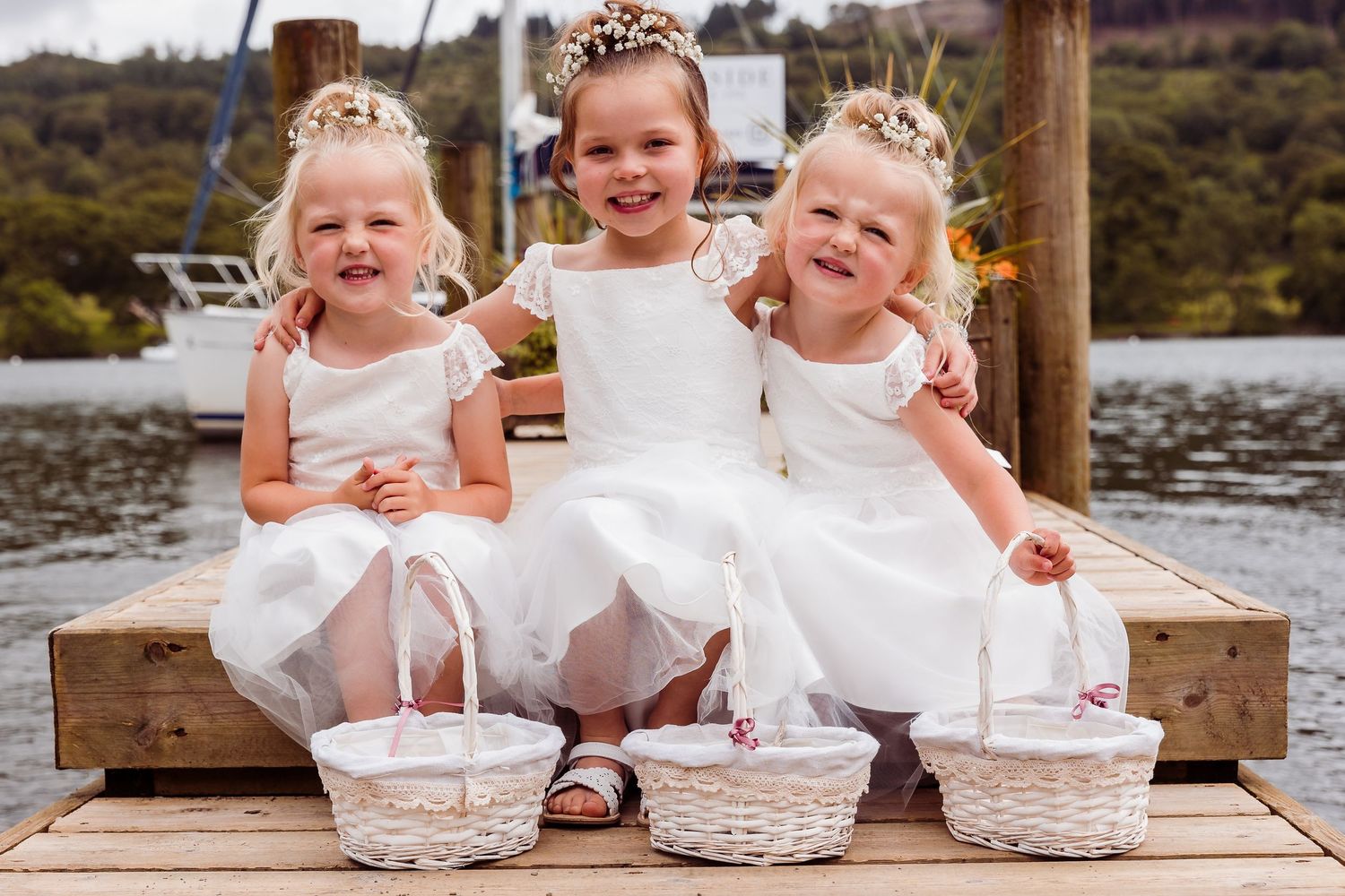 Three flower girls in white dresses sitting on a wooden dock, each holding a white wicker basket.