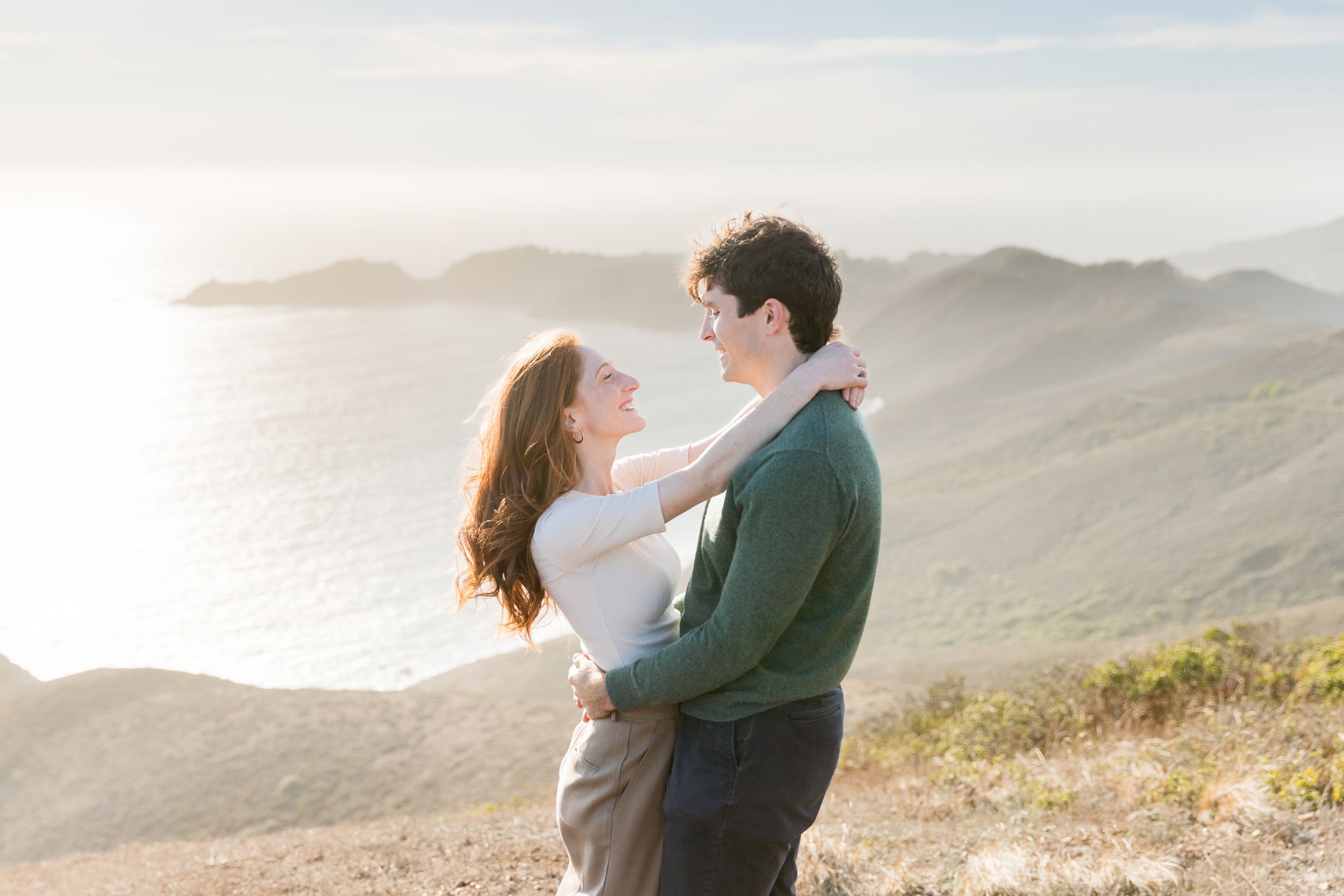Couple embracing on a scenic coastal hillside at golden hour with ocean and mountains in the background.