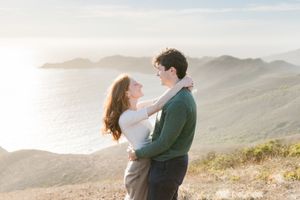 Couple embracing on a scenic coastal hillside at golden hour with ocean and mountains in the background.