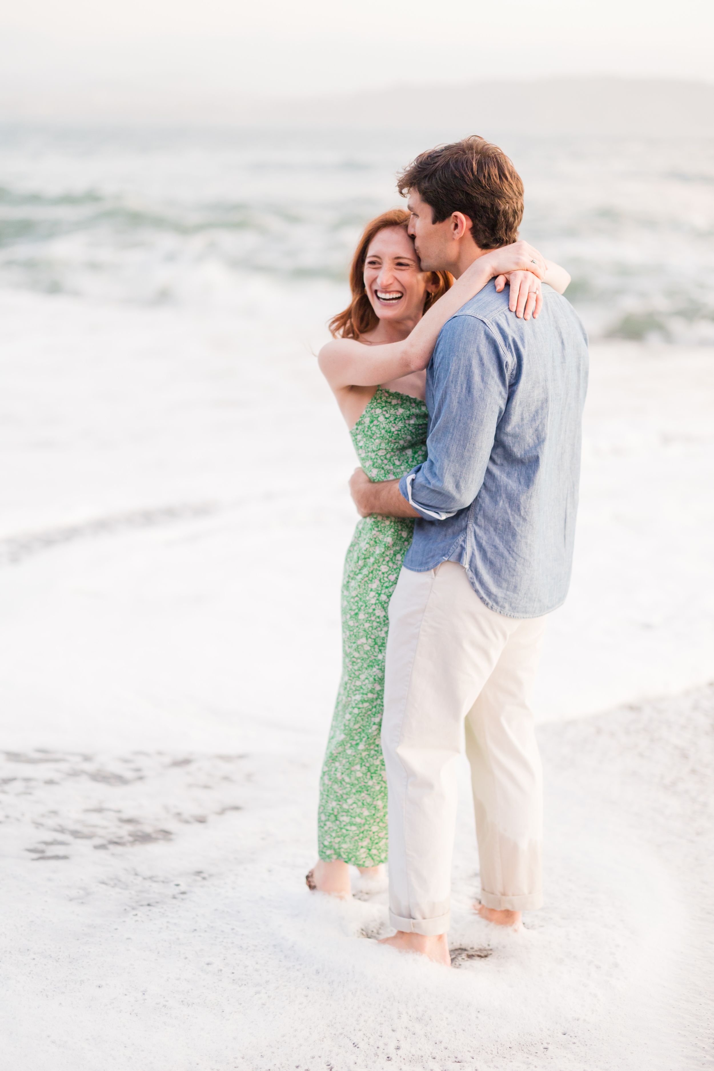 Couple embracing and laughing on a beach, woman in green floral dress, man in blue shirt and white pants.
