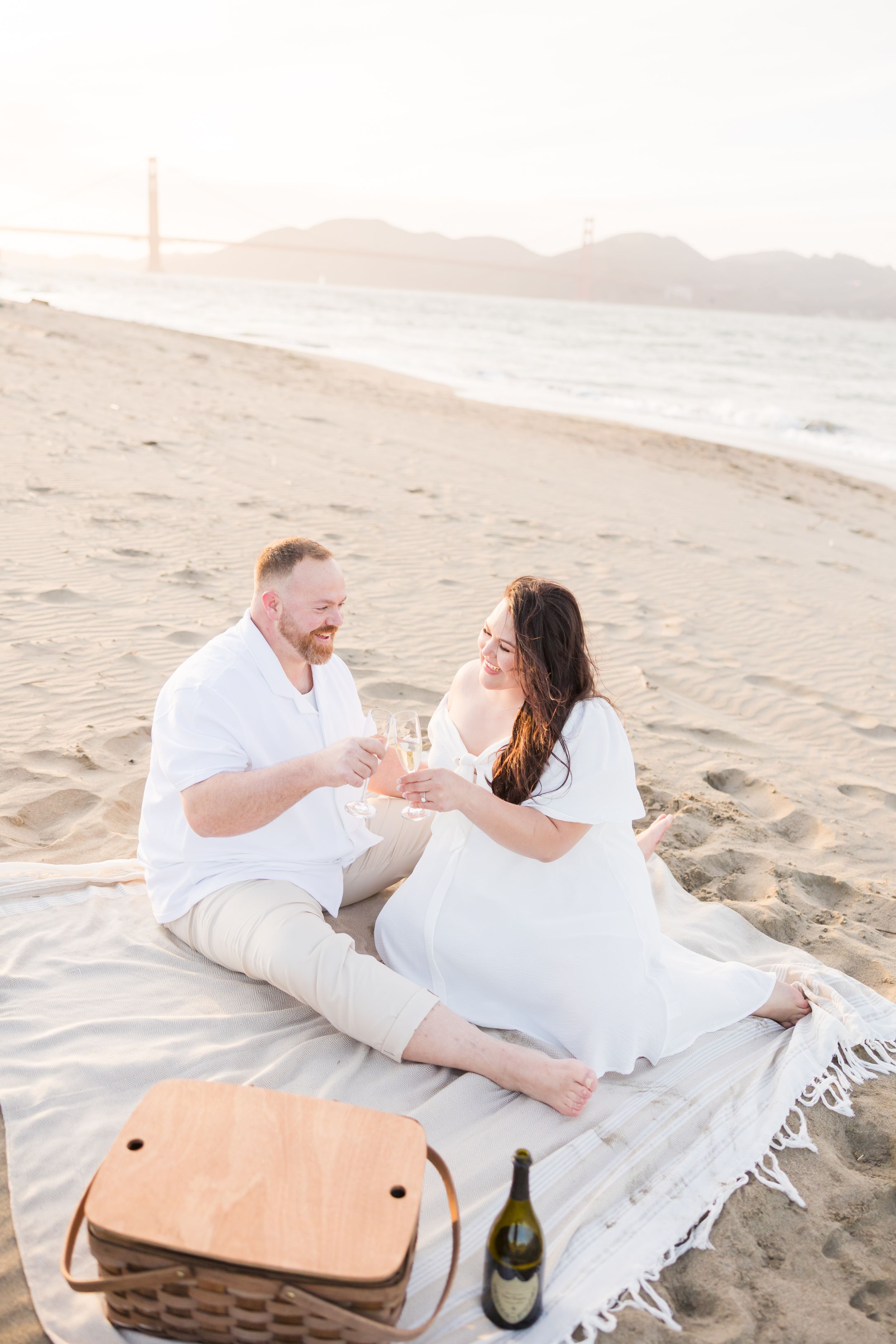 Couple toasting champagne on a beach blanket with Golden Gate Bridge in the background at sunset.
