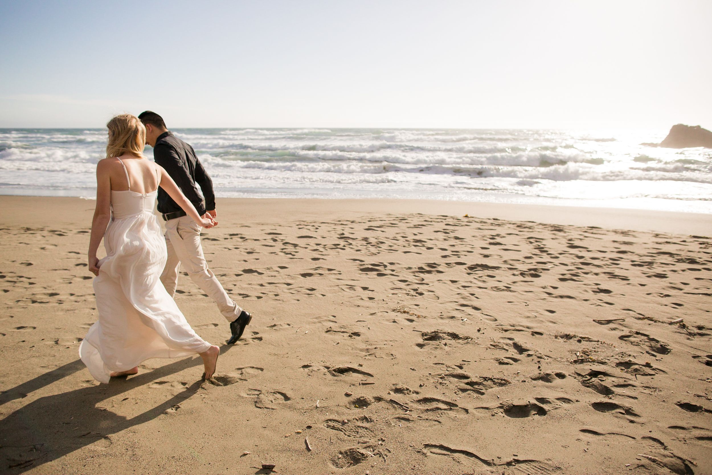 Bride and groom walking hand in hand on a sandy beach near ocean waves on their wedding day.