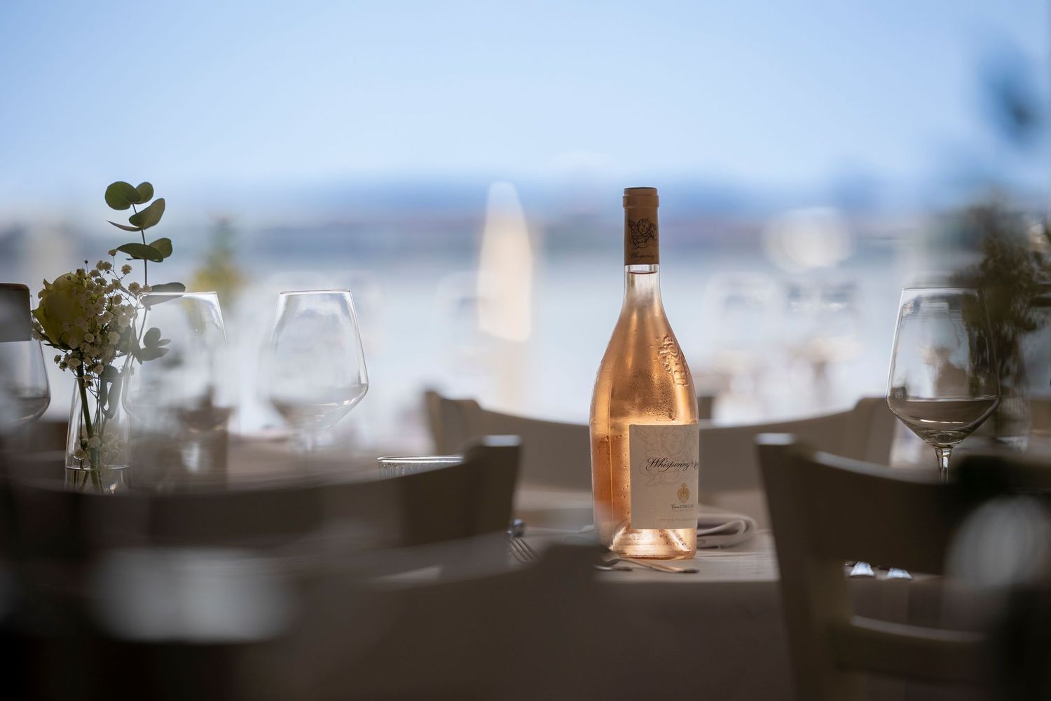 Elegant rosé wine bottle centerpiece on a set dining table with glassware, overlooking a serene waterfront view.