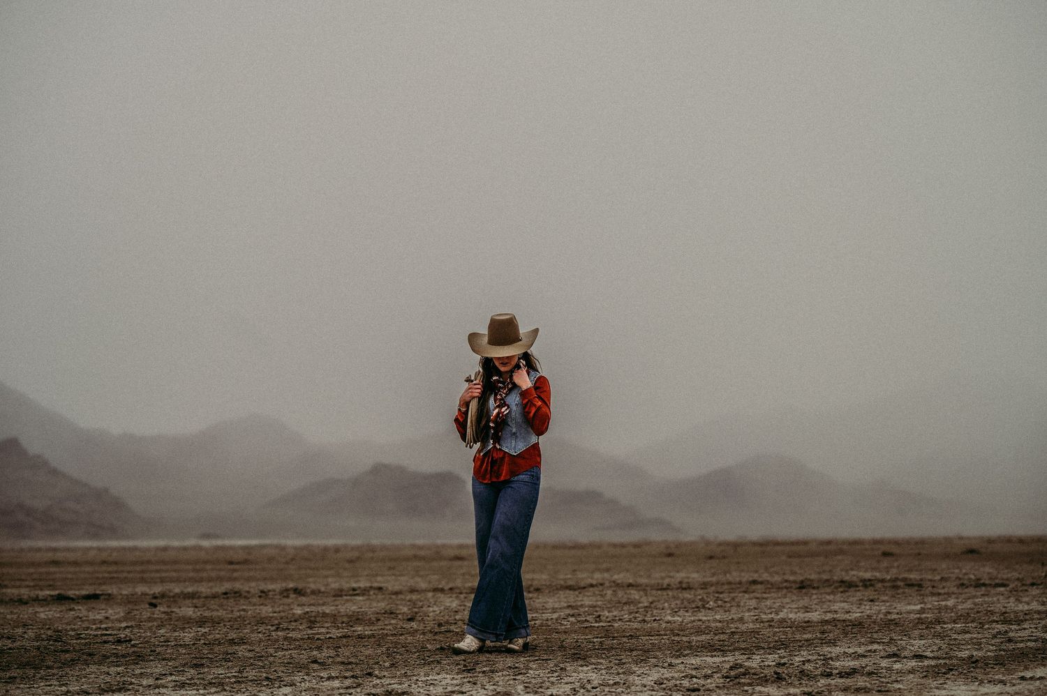 Woman in gray fur coat and brown cowboy hat tilting brim down, wearing turquoise bracelet, snowy mountains behind.