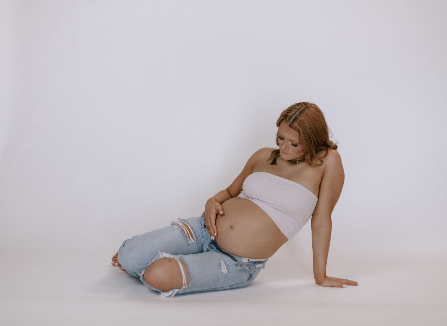 Pregnant woman in pink tube top and ripped jeans sitting on white floor holding her bump.