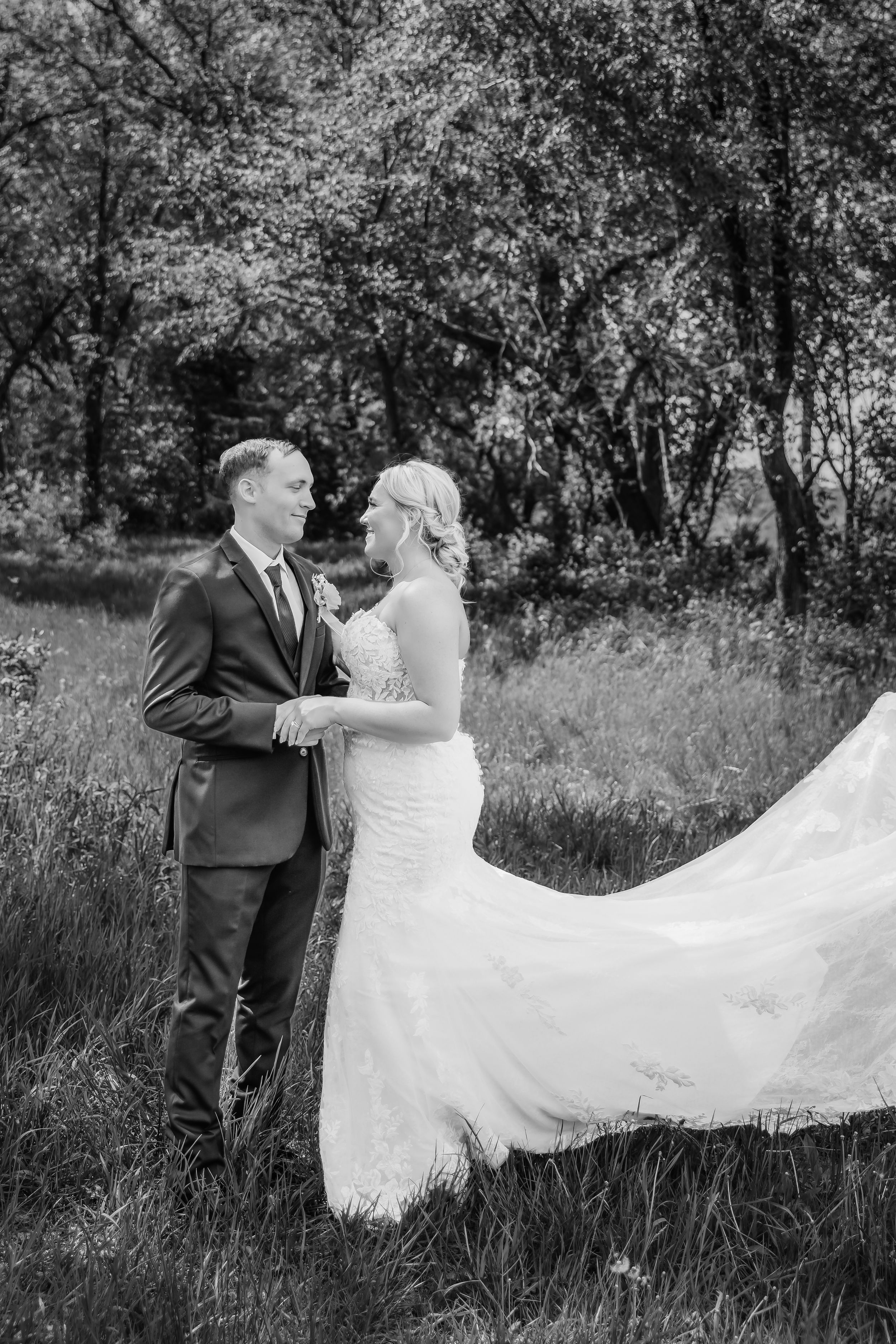 Black and white photo of a bride and groom holding hands in a lush outdoor setting, her gown flowing in the breeze.