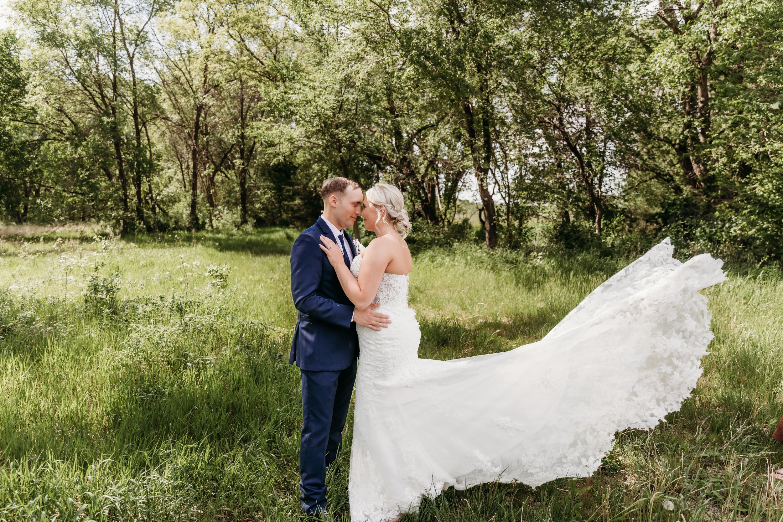 Bride and groom share a romantic moment in a lush green meadow, with the wedding veil flowing in the breeze.