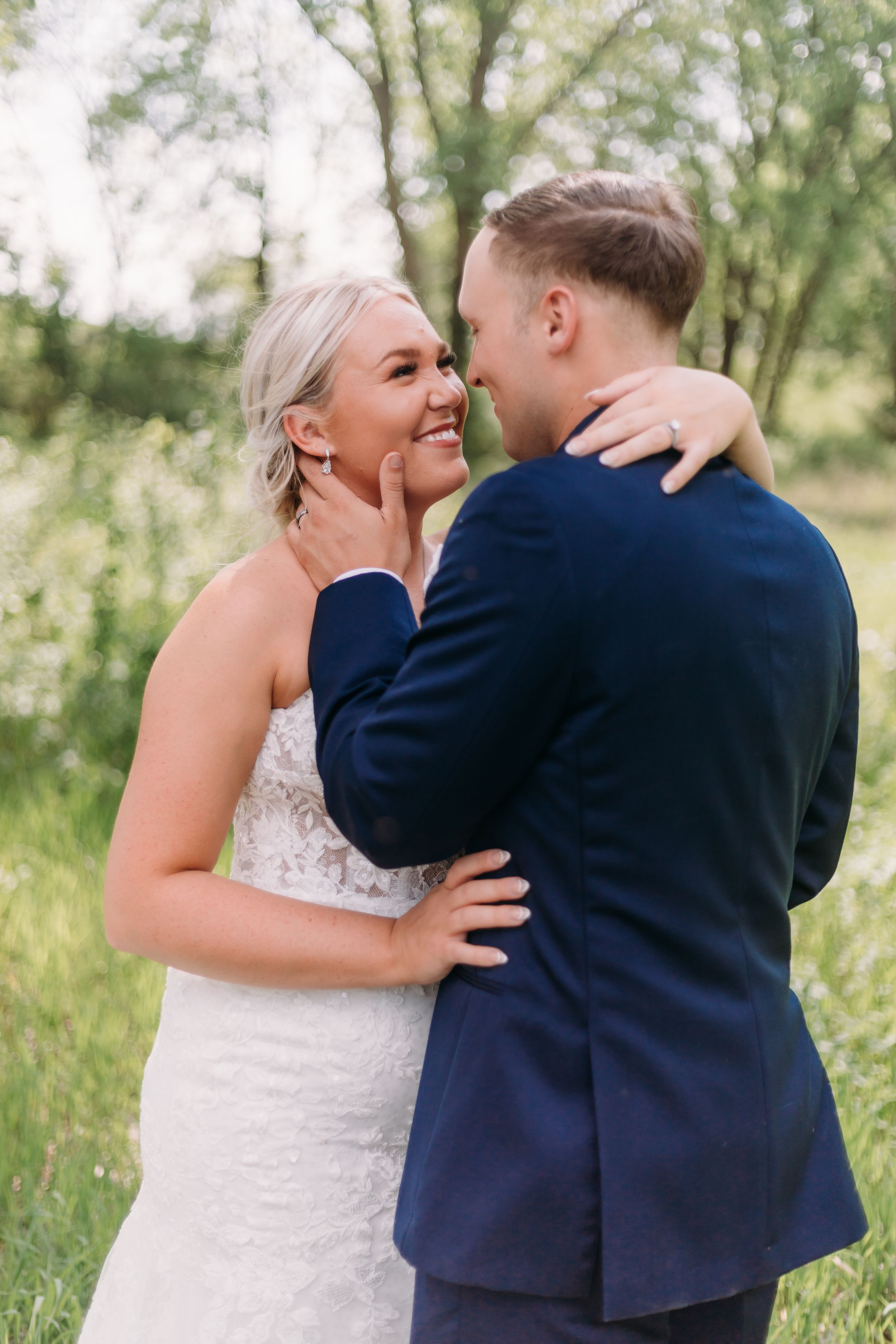 Bride in lace gown smiles as groom in navy suit tenderly cups her face outdoors.
