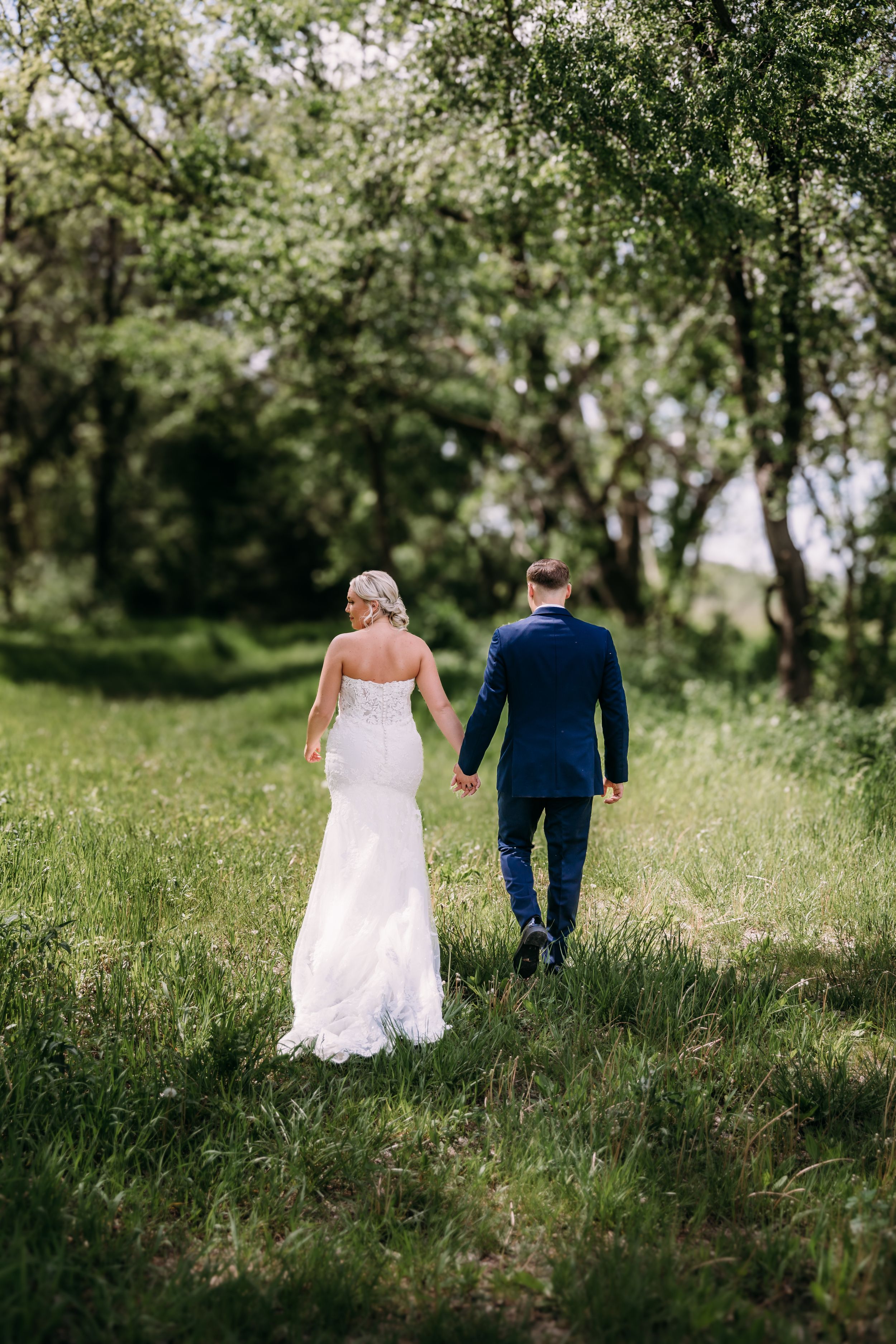 Bride and groom walking hand in hand through lush green meadow, bride in white gown, groom in navy suit.