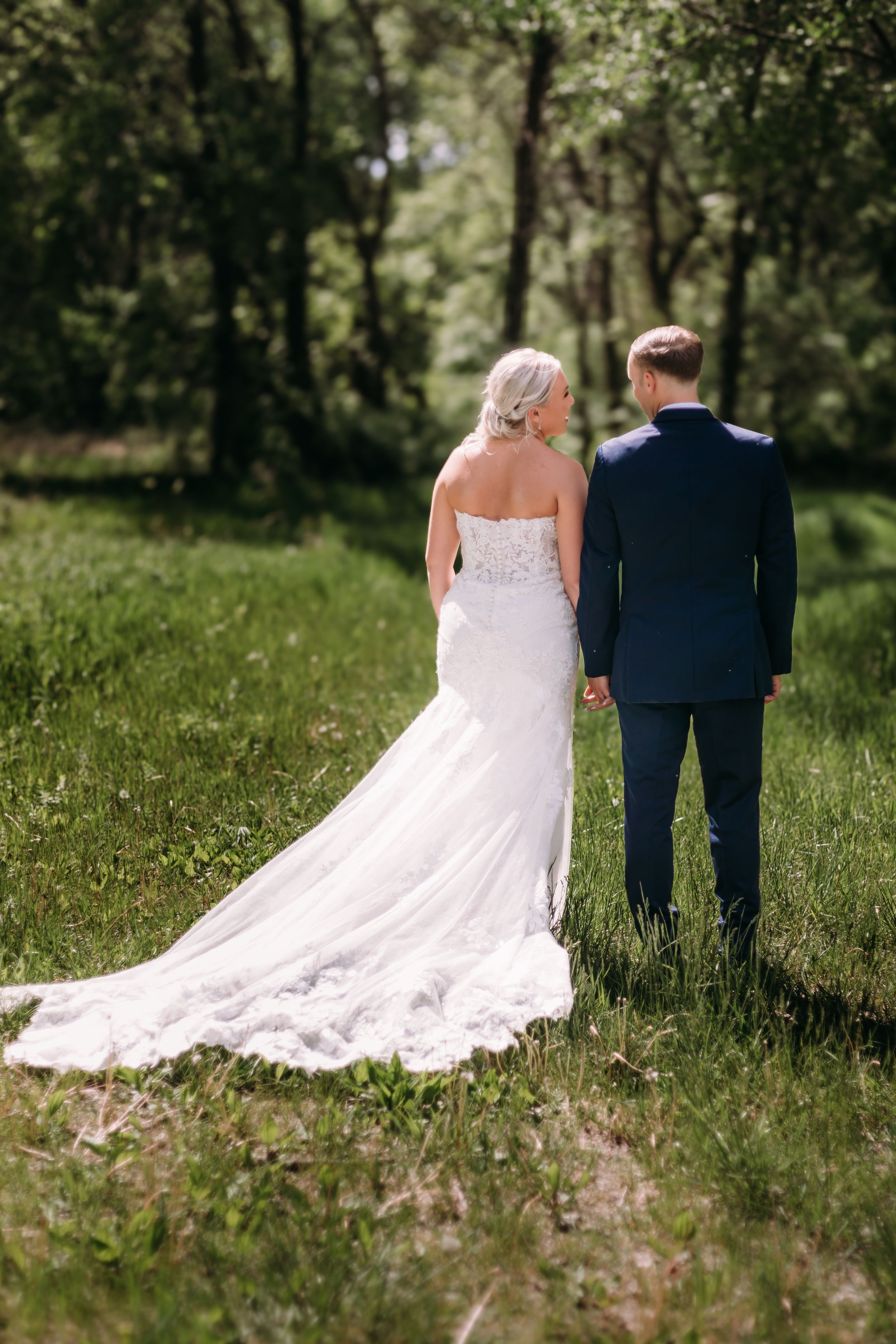 Bride and groom holding hands walking through lush green forest, bride wearing elegant white lace gown with long train.