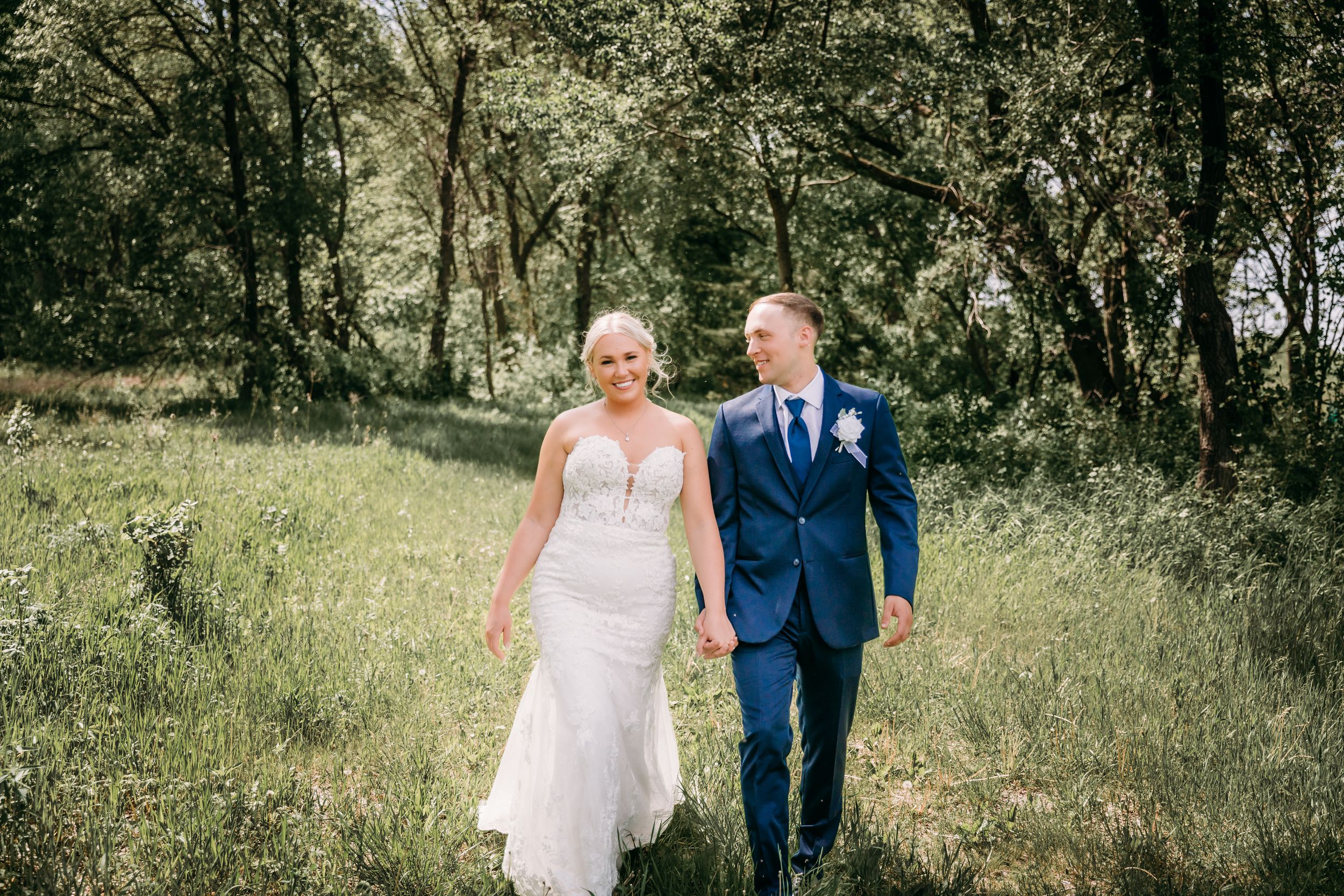 Bride and groom walk hand in hand through a lush green meadow, she in a lace gown, he in a navy suit.