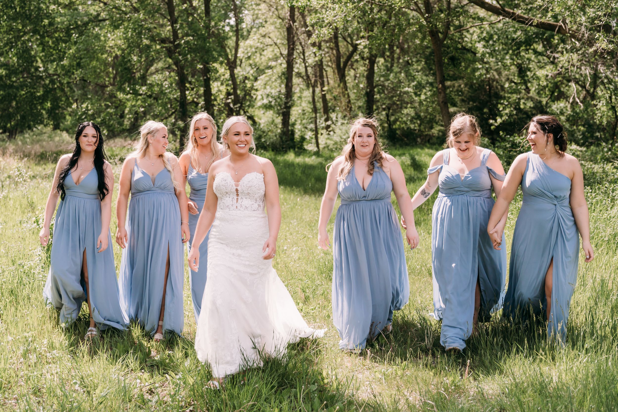 Bride in white gown walks with six bridesmaids in blue dresses through a lush green meadow.