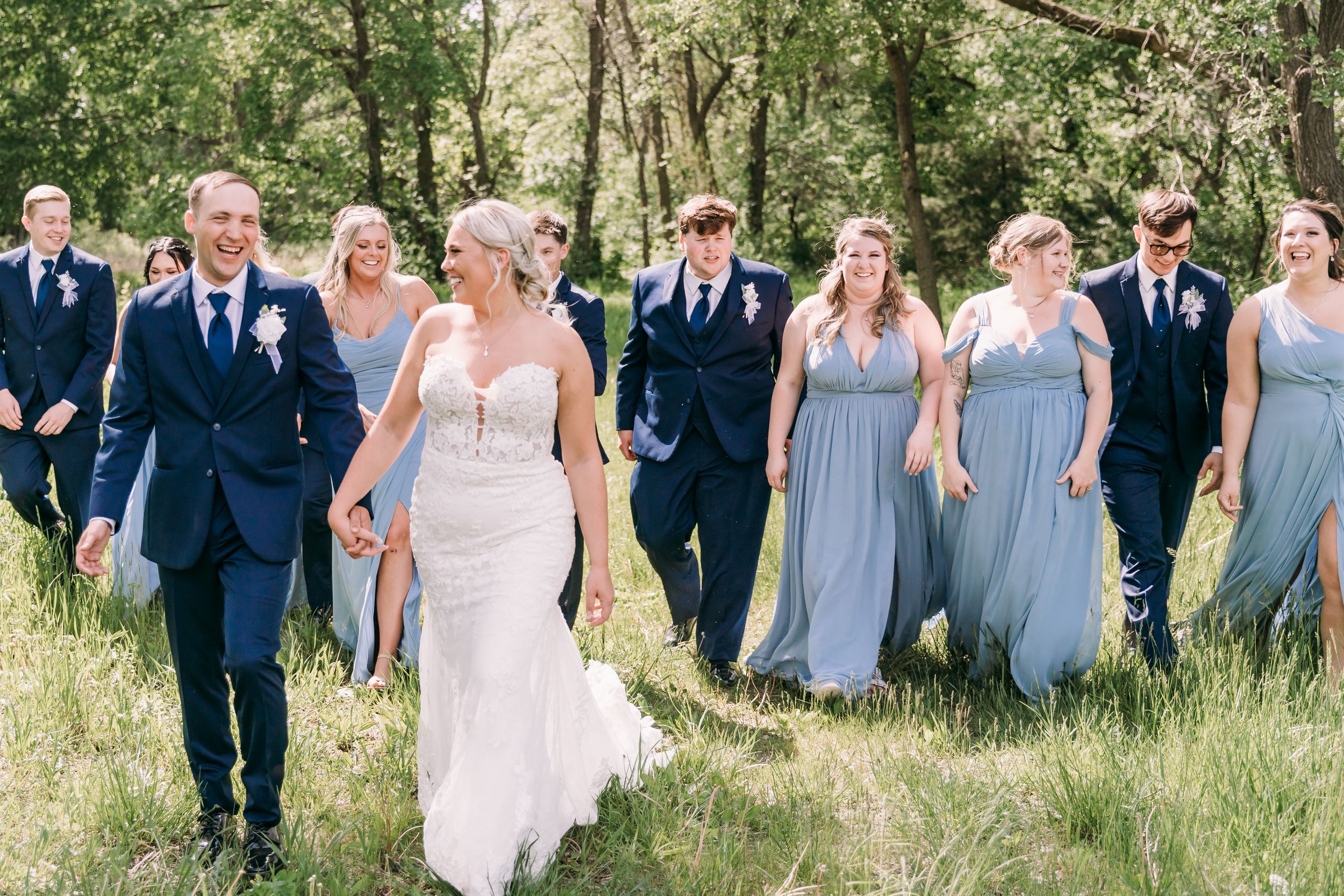 Wedding party walks outdoors in lush green field, bride in white gown with groom in navy suit, bridesmaids in blue dresses.