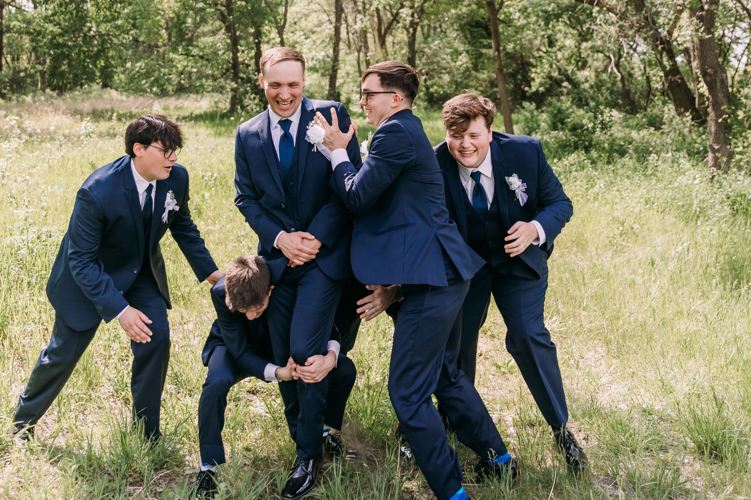 Groomsmen in navy suits playfully posing together outdoors in a green meadow during a wedding celebration.