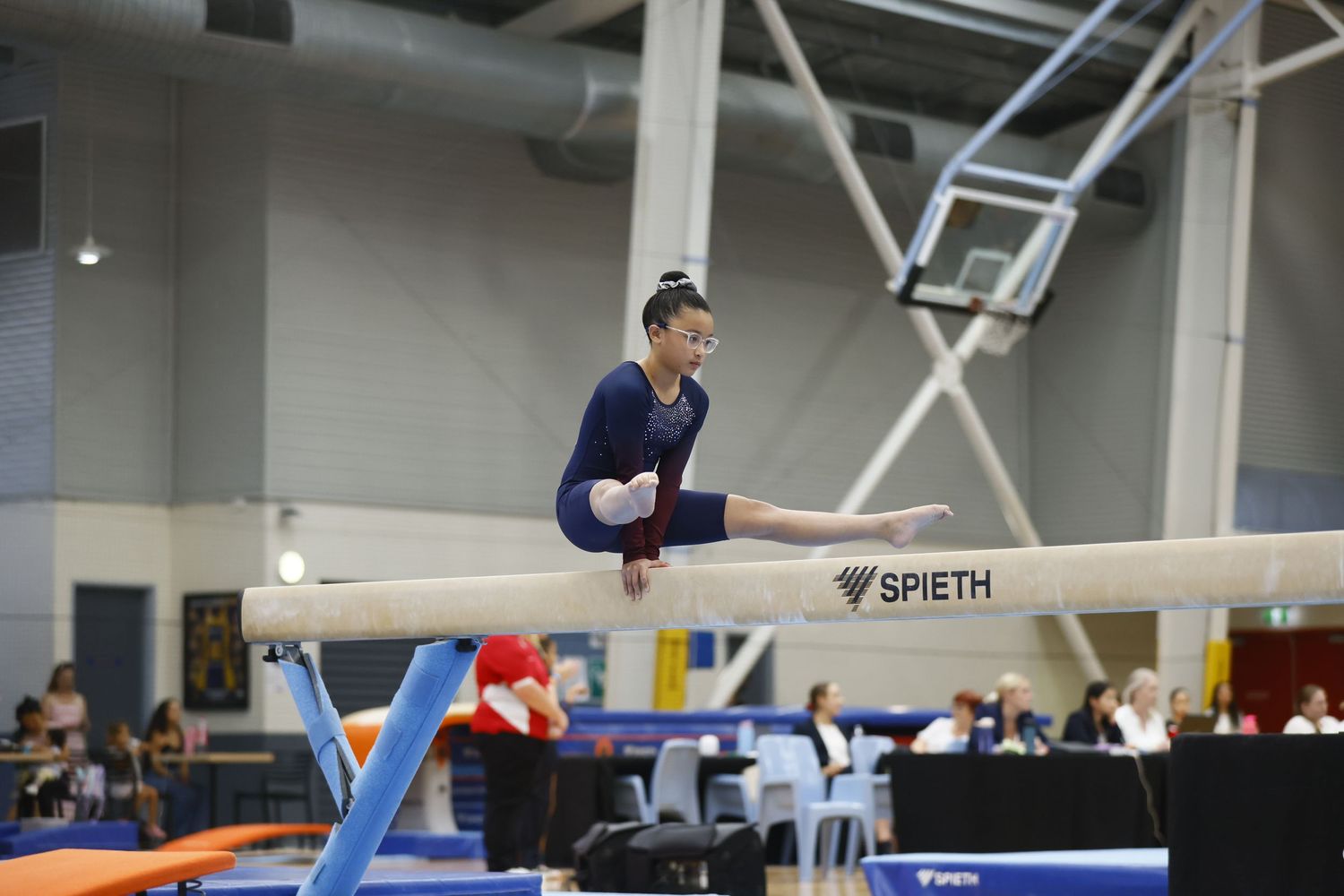 Female gymnast performing on balance beam at indoor gymnastics competition with Spieth equipment visible.