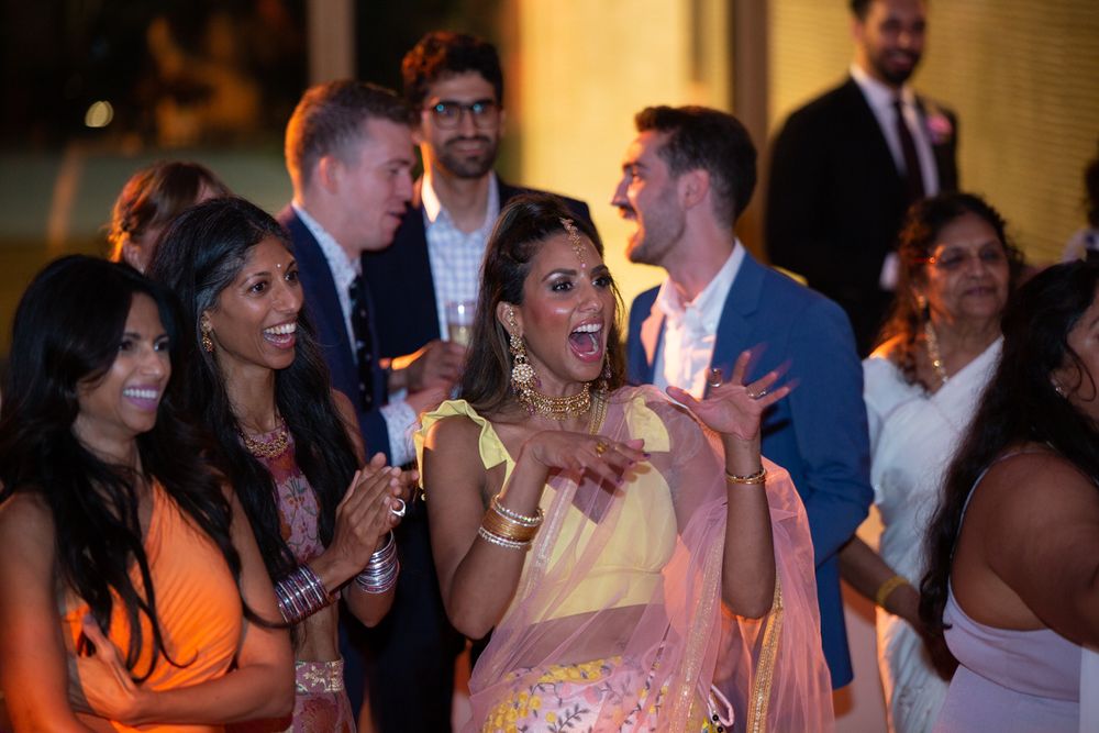 Guests in colorful traditional and formal attire laugh and celebrate at an Indian wedding reception.