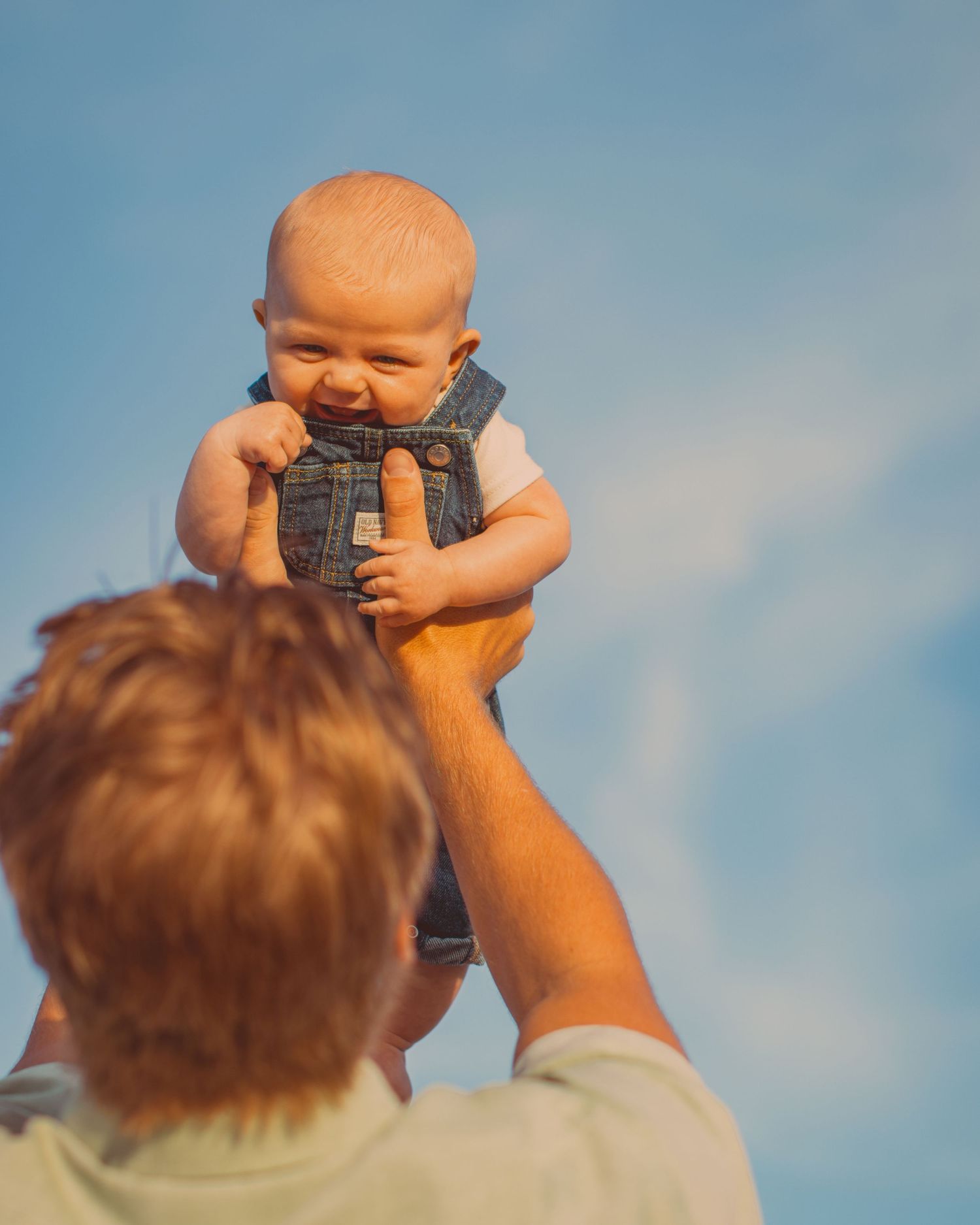Father lifting happy baby in denim vest high into blue sky outdoors.