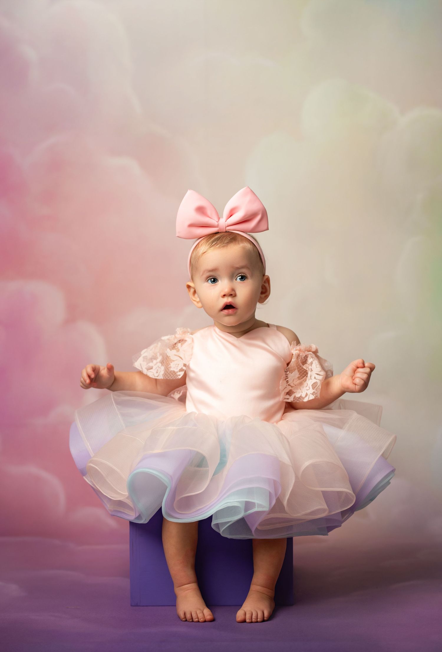 Baby girl in pink tutu dress and bow headband sits on purple block against pastel cloud background.
