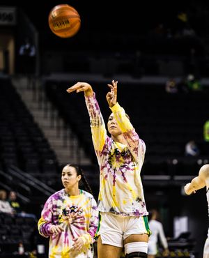 Women in tie-dye shirts practice basketball drills on an indoor court, one player shooting the ball.