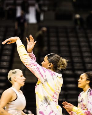 Women in tie-dye outfits on a basketball court, one raising her hand, with empty stadium seats in the background.