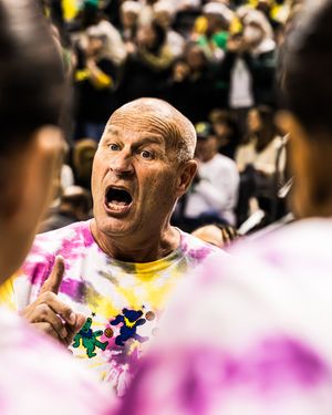 Bald man in tie-dye shirt yelling intensely at players during a sports event with crowd in background.