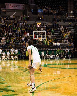 A basketball player stands on a green court during a college game in a packed arena with fans in the stands.