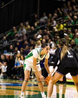Women's college basketball player in green uniform dribbles against defender wearing number 1 jersey during indoor game.