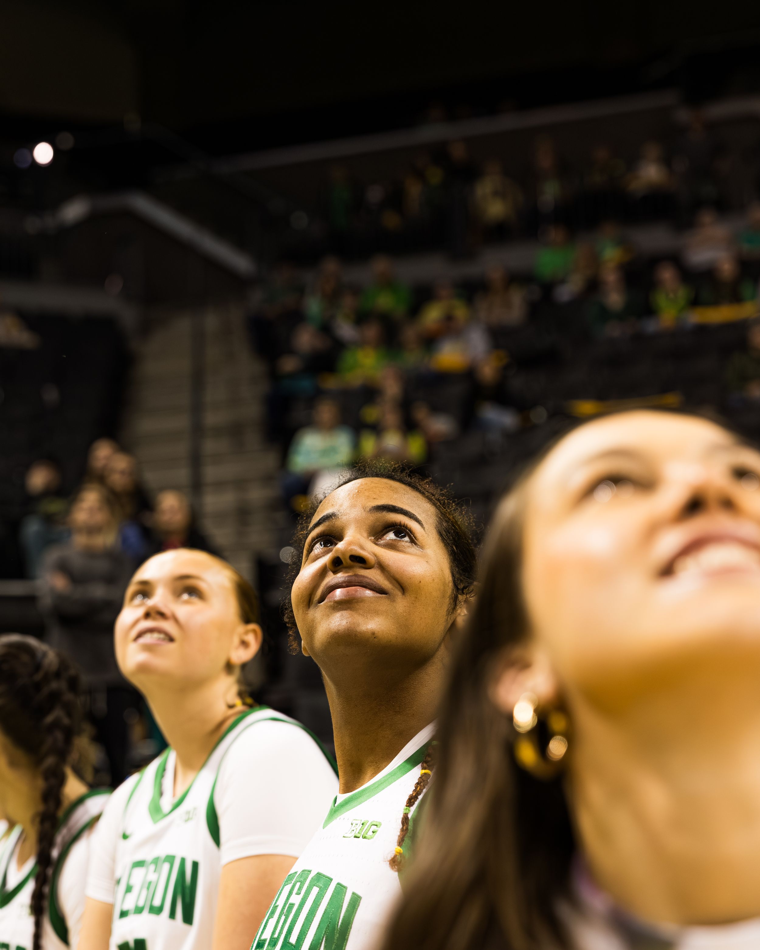 Oregon Ducks women's basketball players in white uniforms look up during a game at a packed indoor arena.