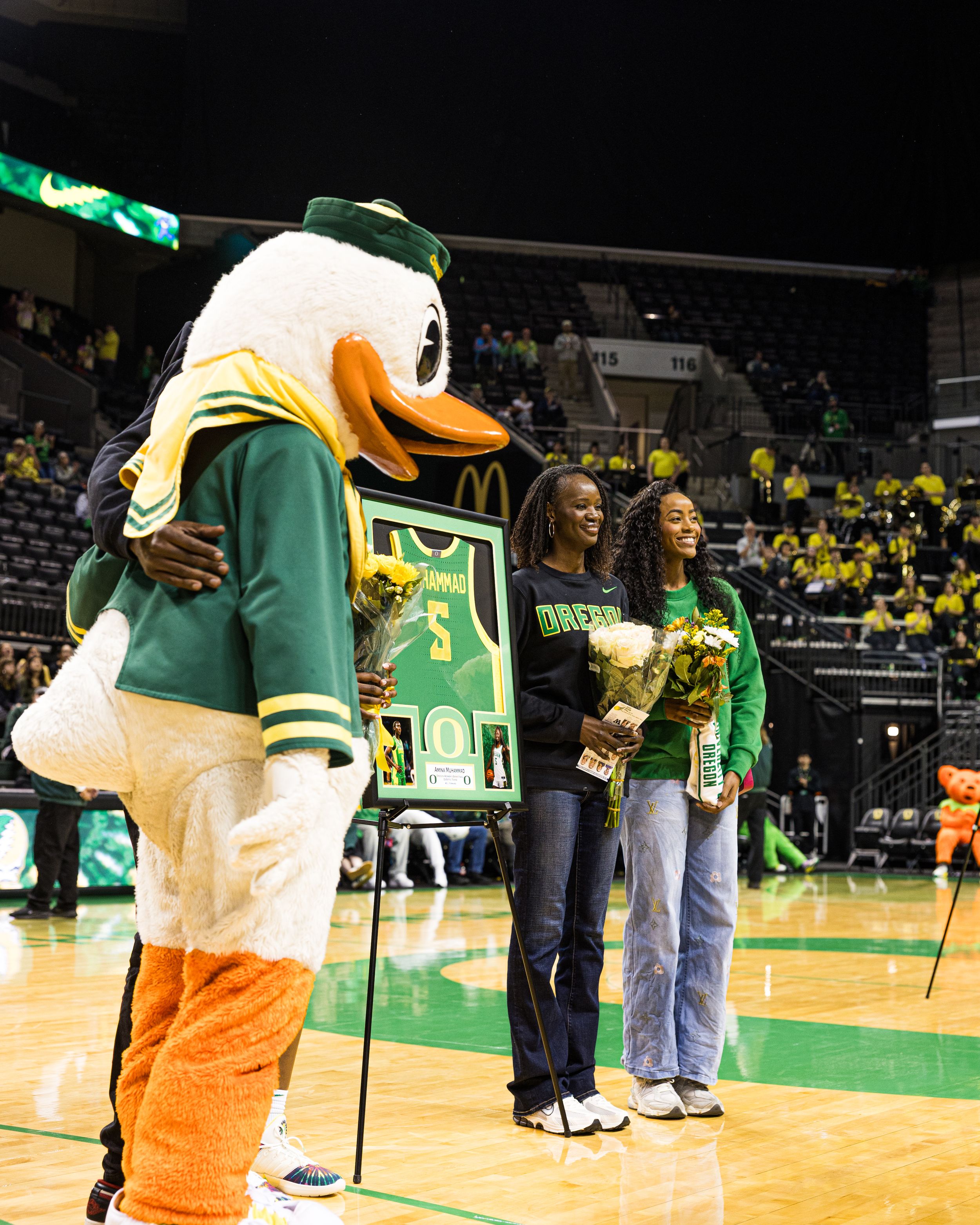 Oregon Ducks mascot poses with two students holding trophies during a basketball game ceremony at Matthew Knight Arena.
