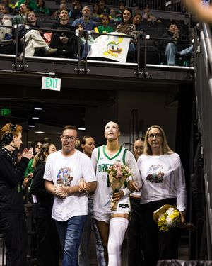 Oregon Ducks basketball player in green jersey holds flowers with family during senior night ceremony at arena.