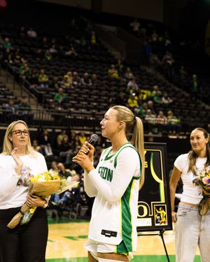 Female basketball player speaks at microphone during senior day ceremony on indoor court with flowers and teammates nearby.