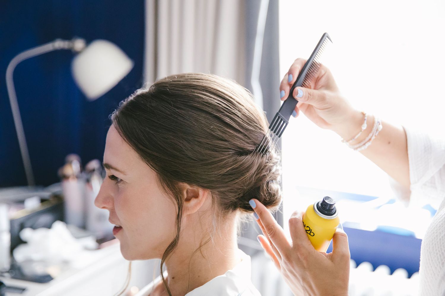 A hairstylist applying hairspray while styling a brunette woman's elegant updo for a wedding or special event.