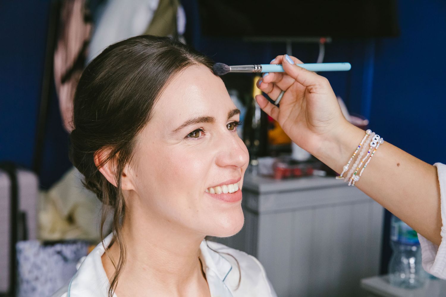 A smiling bride gets her makeup applied with a brush by a makeup artist during wedding preparations.