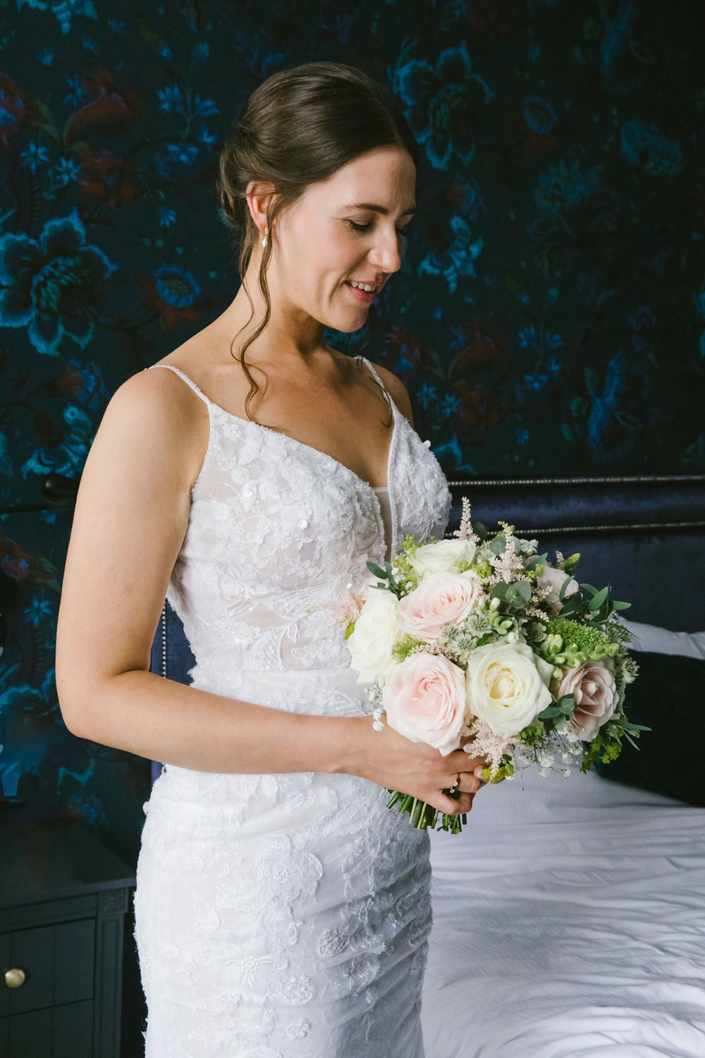 Smiling bride in lace wedding gown holding a bouquet of blush pink and white roses in an elegant bedroom.
