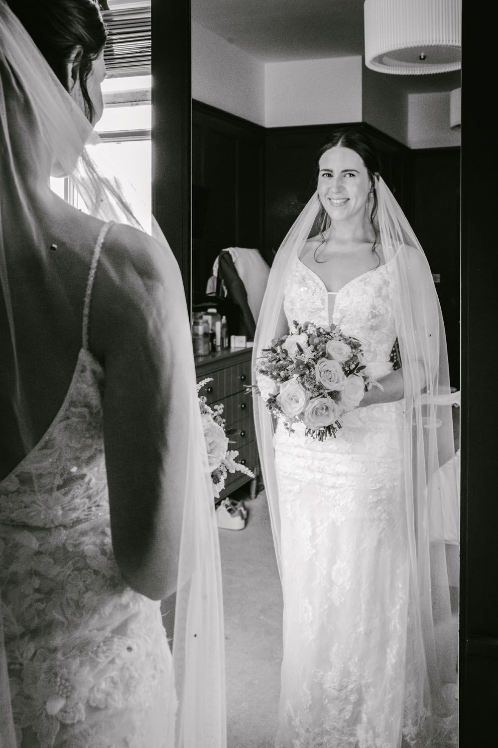 Black and white photo of a smiling bride in lace wedding dress and veil holding bouquet, reflected in mirror.