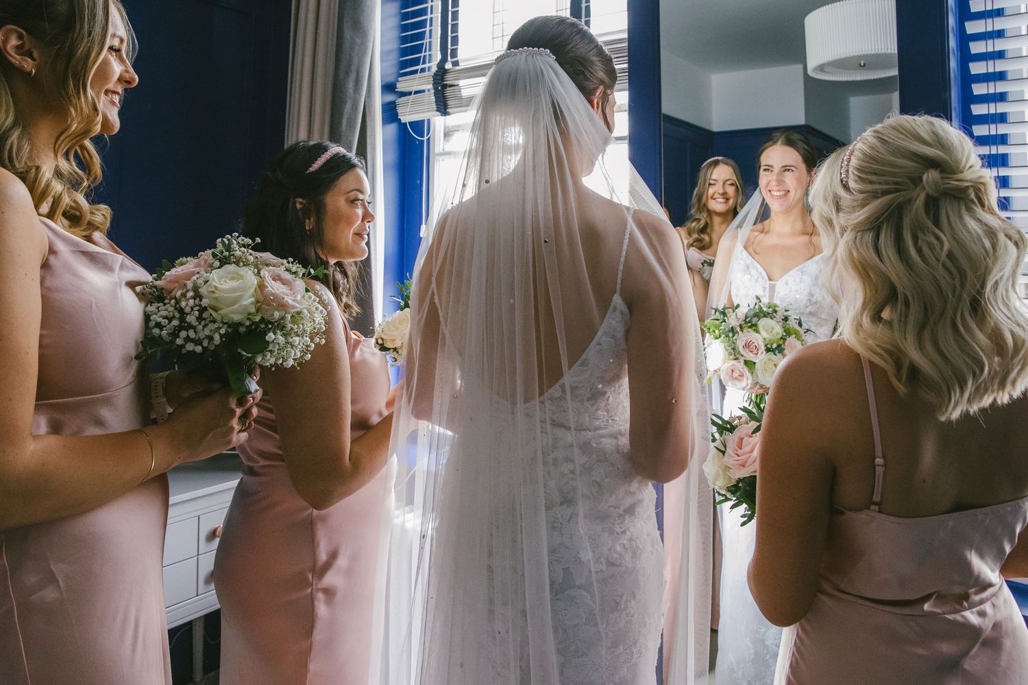 Bride in white lace gown and veil surrounded by bridesmaids in pink dresses holding bouquets in elegant blue room.